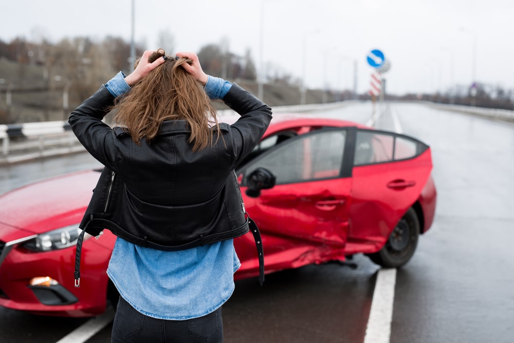 Woman Holding Her Head In Shock Next To A Crashed Car