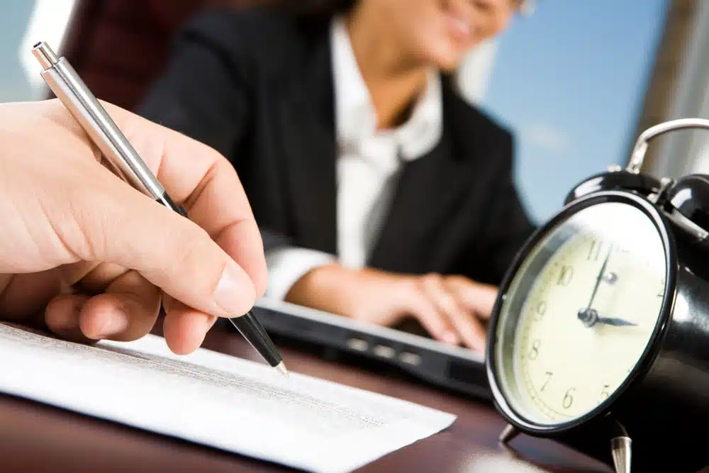 Close-up of male's hand with pen over document on background of working woman